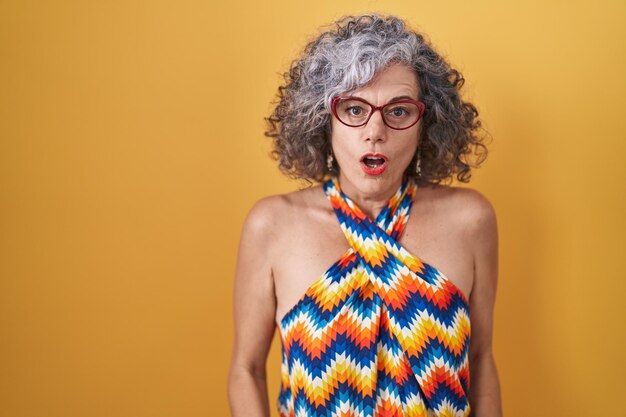 Middle age woman with grey hair standing over yellow background afraid and shocked with surprise and amazed expression, fear and excited face.