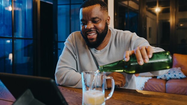 Афро-американский мужчина средних лет пьет пиво и веселится через видеозвонок в гостиной дома.
