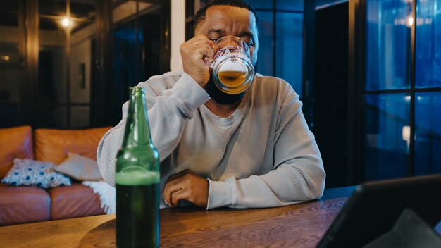 Middle-aged African American male drinking beer having fun happy night party event online celebration via video call in living room at home.