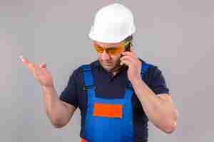 Бесплатное фото Злой строитель средних лет в строительной форме и защитном шлеме разговаривает по мобильному телефону, раздраженный и разочарованный из-за изолированной белой стены