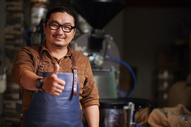 Азиатский мужчина средних лет позирует с большим пальцем перед оборудованием для обжарки кофе