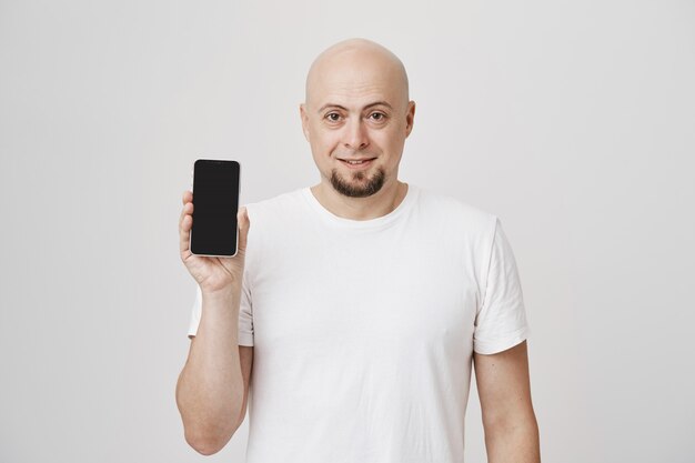 Лысый бородатый парень средних лет в белой футболке показывает приложение для смартфона