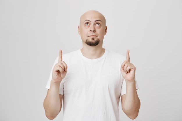 Middle-aged bald bearded man in white t-shirt looking and pointing up at copyspace