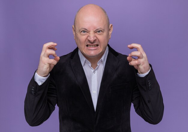 Middle-aged bald man in suit going wild raising arms yelling with aggressive expression standing over purple wall