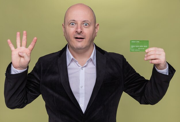 Middle-aged bald man in suit holding credit card looking at front smiling surprised showing number four with fingers standing over green wall