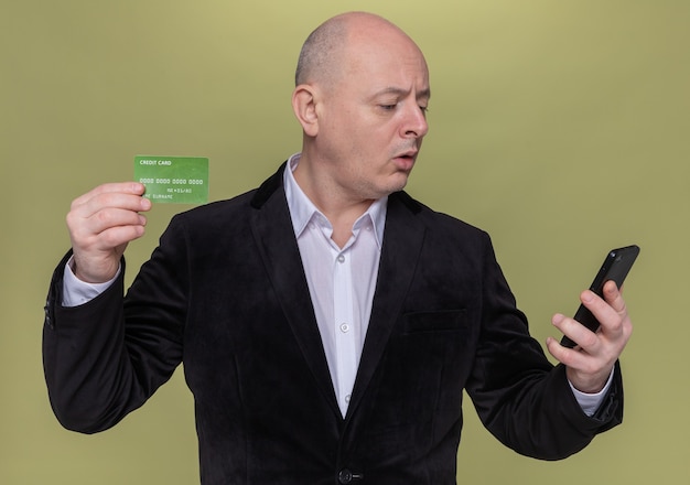 Uomo calvo di mezza età in vestito che tiene la carta di credito guardando il telefono cellulare confuso in piedi sopra la parete verde