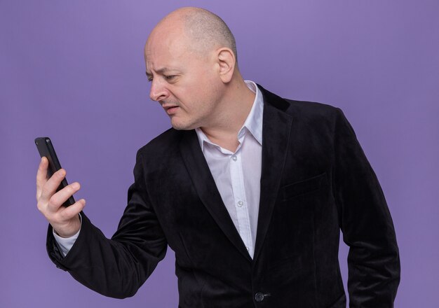 Middle-aged bald man in suit holding mobile phone looking at it intrigued standing over purple wall