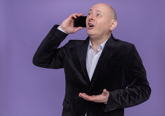 Middle-aged bald man in suit looking surprised and happy while talking on mobile phone standing over purple wall