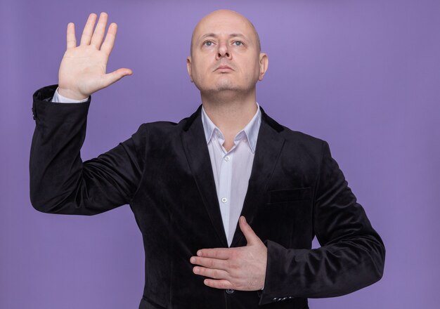Middle-aged bald man in suit making a promise holding hand on chest and raising other hand standing over purple wall
