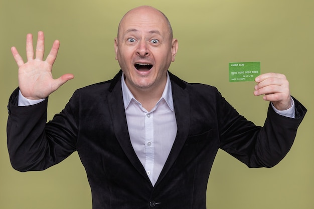 Foto gratuita uomo calvo di mezza età in vestito che mostra la carta di credito che è felice ed eccitato che mostra la palma aperta numero cinque che sta sopra la parete verde