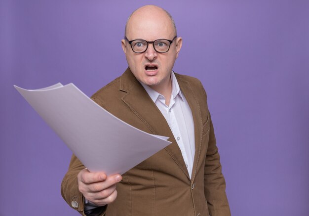 Middle-aged bald man in suit wearing glasses holding blank pages looking aside with aggressive expression standing over purple wall