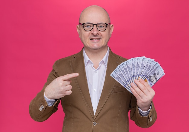 Middle-aged bald man in suit wearing glasses holding cash looking at front pointing with index finger at money happy and positive standing over pink wall