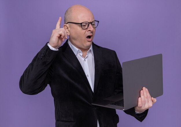 Лысый мужчина средних лет в костюме в очках, держащий ноутбук, выглядит удивленным, показывая указательный палец с новой идеей, стоящим над фиолетовой стеной
