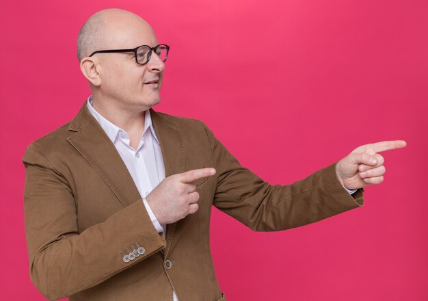 Middle-aged bald man in suit wearing glasses looking aside smiling confident pointing with index fingers to the side standing over pink wall