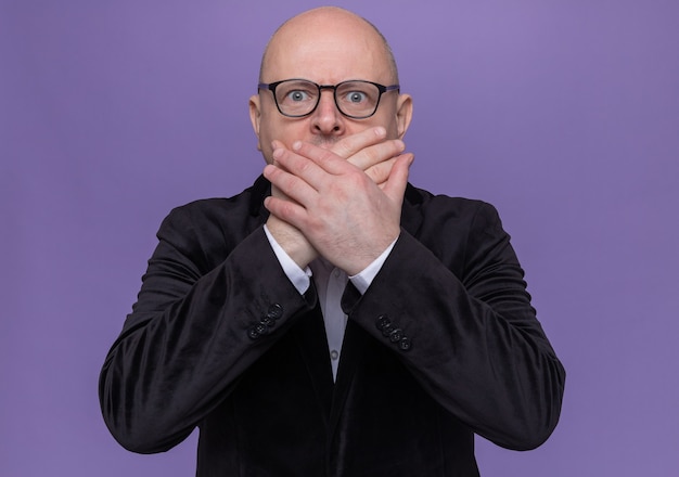 Free photo middle-aged bald man in suit wearing glasses looking at front bein shocked covering mouth with hands standing over purple wall