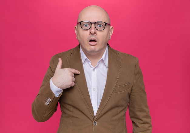 Middle-aged bald man in suit wearing glasses looking at front surprised pointing at himself standing over pink wall