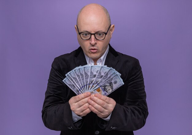 Middle-aged bald man in suit wearing glasses looking at money happy and excited standing