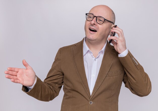 Middle-aged bald man in suit wearing glasses smiling broadly happy and cheerful while talking on mobile phone standing over white wall