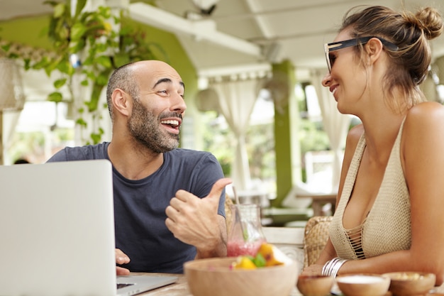 Middle-aged bearded man with cheerful expression pointing thumb at his girlfriend in sunglasses while telling jokes.