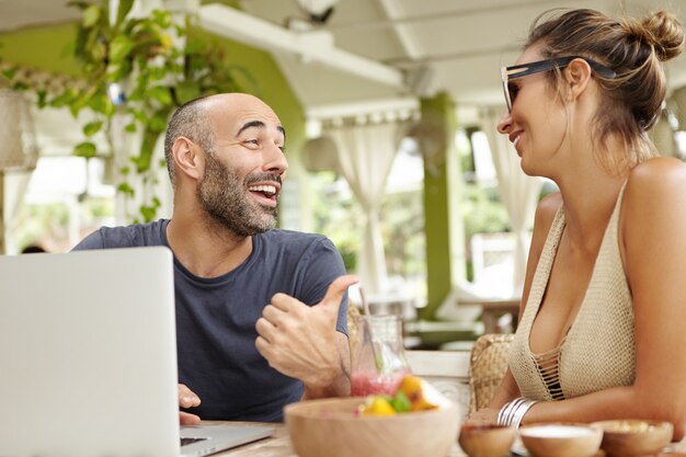 Middle-aged bearded man with cheerful expression pointing thumb at his girlfriend in sunglasses while telling jokes.