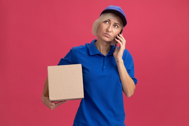 Middle-aged blonde delivery woman in blue uniform and cap holding cardboard box talking on phone looking up 