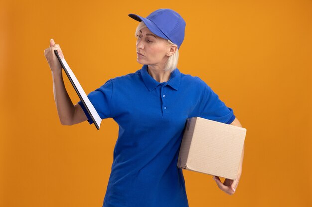 Middle-aged blonde delivery woman in blue uniform and cap holding cardbox underarm and clipboard looking at clipboard 