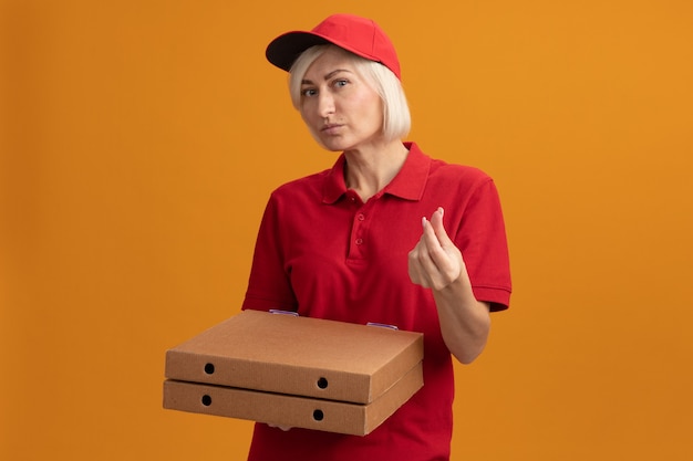 Middle-aged blonde delivery woman in red uniform and cap holding pizza packages  doing money gesture isolated on orange wall with copy space