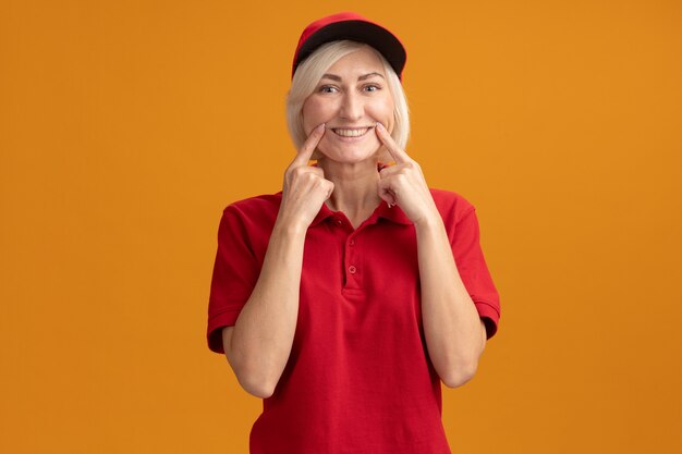 Среднего возраста блондинка доставщица в красной форме и кепке смотрит вперед, делая фальшивую улыбку, изолированную на оранжевой стене с копией пространства