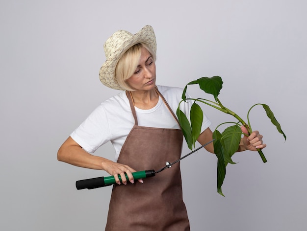 Бесплатное фото Светловолосая женщина-садовник средних лет в униформе в шляпе смотрит на растение, срезающее его ножницами для живой изгороди