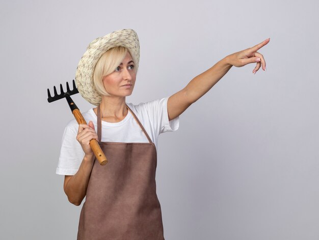 Middle-aged blonde gardener woman in uniform wearing hat holding rake on shoulder looking and pointing up isolated on white wall