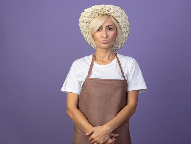 Middle-aged blonde gardener woman in uniform wearing hat keeping hands together 