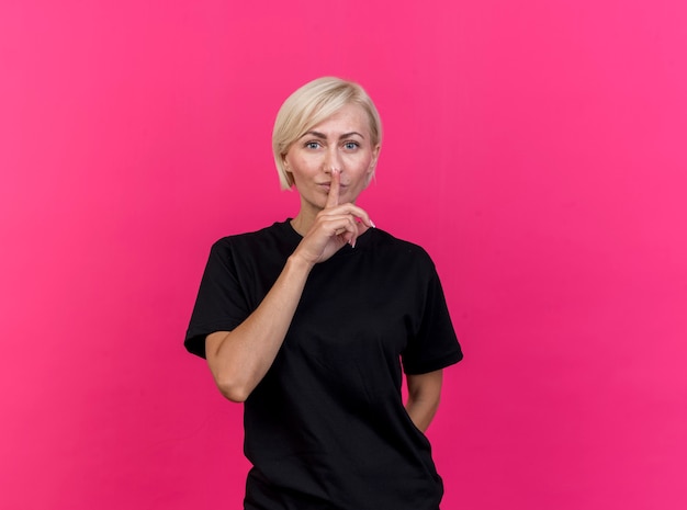 Middle-aged blonde slavic woman looking at camera keeping hand behind back doing silence gesture isolated on crimson background with copy space