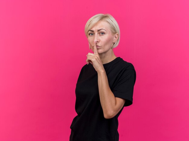 Middle-aged blonde slavic woman standing in profile view  doing silence gesture isolated on crimson wall with copy space