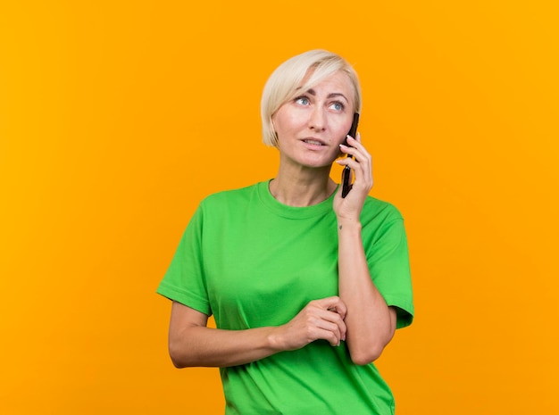 Middle-aged blonde slavic woman talking on phone looking at side isolated on yellow wall with copy space