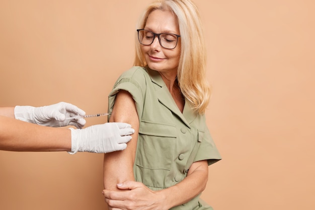 中年の金髪の女性が注射器を見て、ベージュの壁に分離されたパンデミックの間にコロナウイルスに対するワクチンを取得します