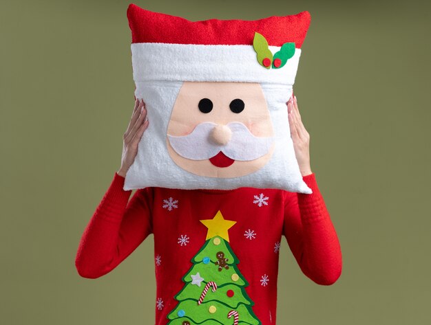 サンタクロースのヘッドバンドとオリーブグリーンの背景で隔離の顔の前にサンタクロースの枕を保持しているクリスマスセーターを着ている中年のブロンドの女性