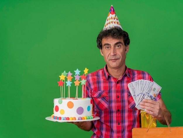 Free photo middle-aged caucasian party man wearing birthday cap holding birthday cake paper bag gift pack and money looking at camera isolated on green background with copy space