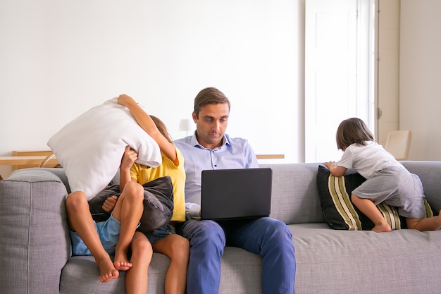 Middle-aged dad sitting on sofa and working on laptop when kids playing near him. Caucasian father using computer and children having fun on coach in room. Digital technology and fatherhood concept
