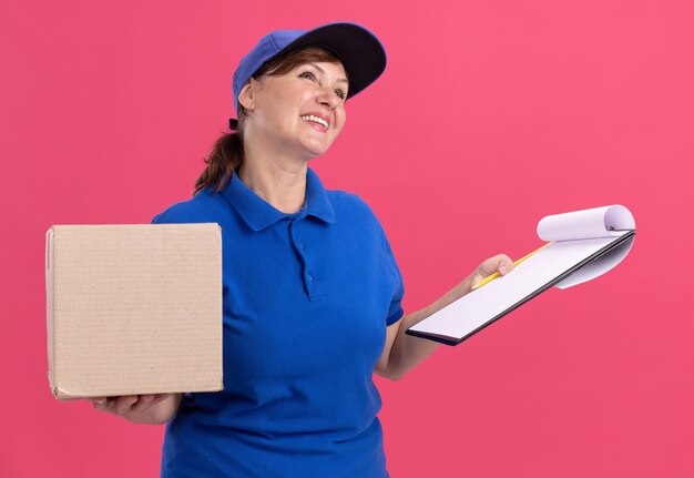 Middle aged delivery woman in blue uniform and cap holding cardboard box and clipboard with blank pages looking up with smile on face standing over pink wall