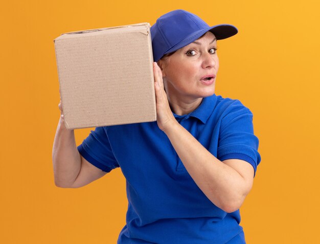 Middle aged delivery woman in blue uniform and cap holding cardboard box over her ear listening standing over orange wall