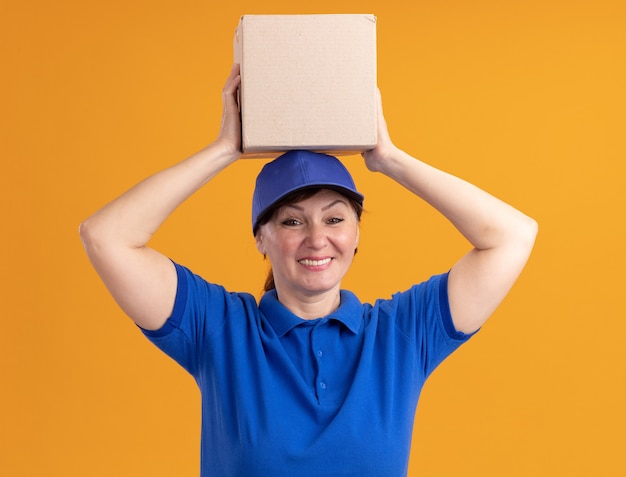 Middle aged delivery woman in blue uniform and cap holding cardboard box over her head looking at front smiling cheerfully standing over orange wall