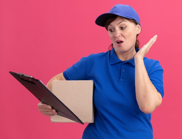 ピンクの壁の上に立って混乱しているクリップボードを見て段ボール箱を保持している青い制服と帽子の中年配達の女性