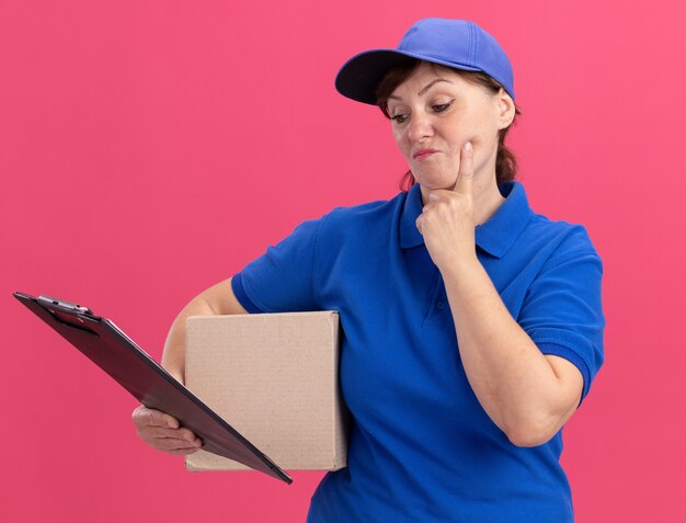 Женщина-доставщик средних лет в синей форме и кепке держит картонную коробку, глядя в буфер обмена с серьезным лицом, стоящим над розовой стеной