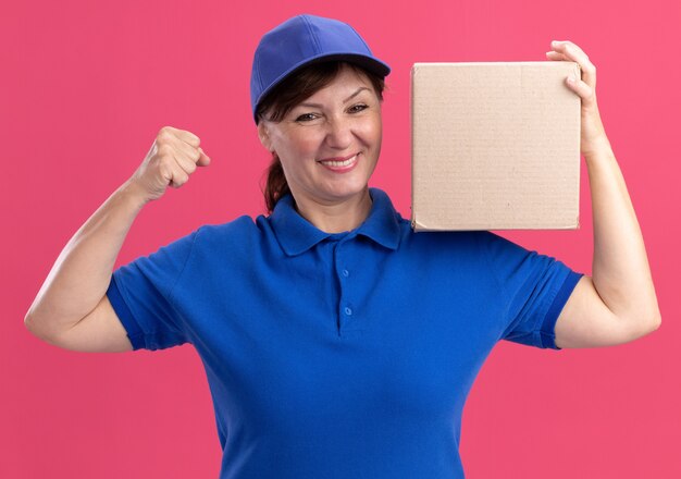 Middle aged delivery woman in blue uniform and cap holding cardboard box looking at front happy and excited clenching fist standing over pink wall