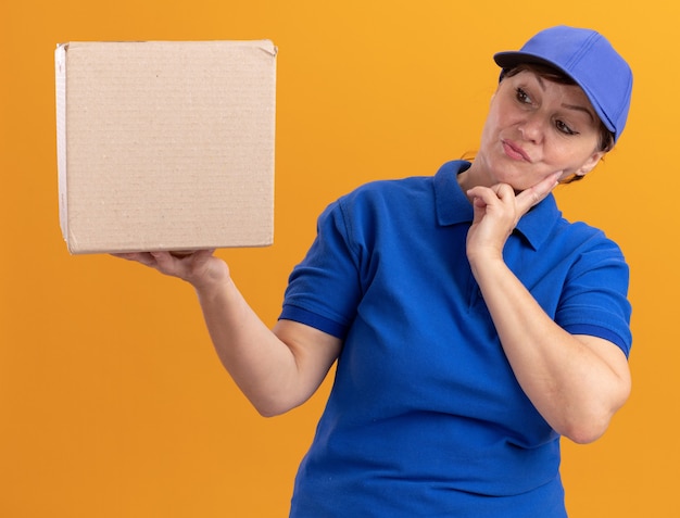 Middle aged delivery woman in blue uniform and cap holding cardboard box looking at it puzzled standing over orange wall