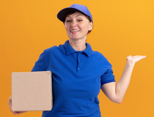 青いユニフォームとオレンジ色の壁の上に立っている幸せそうな顔で笑顔で正面を見て腕で何かを提示する段ボール箱を保持している中年の配達の女性