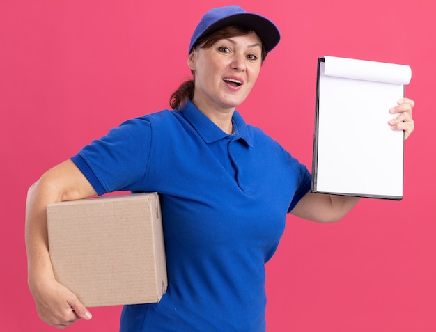 Middle aged delivery woman in blue uniform and cap holding cardboard box showing clipboard with blank pages looking at front happy and positive standing over pink wall