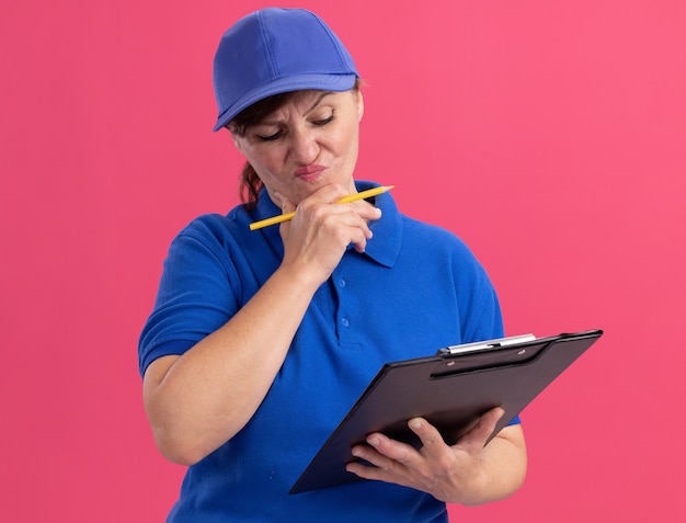 Donna di mezza età delle consegne in uniforme blu e cappuccio che tiene appunti e matita che sembra confusa e molto ansiosa in piedi sopra il muro rosa