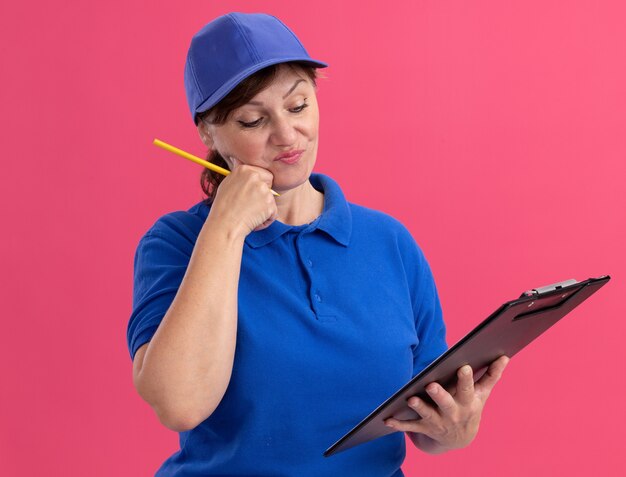 Женщина-доставщик средних лет в синей форме и кепке держит в руках буфер обмена и карандаш, глядя на него с растерянным выражением лица, стоя над розовой стеной