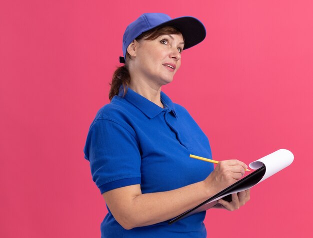Middle aged delivery woman in blue uniform and cap holding clipboard and pencil writing looking with pensive expression thinking standing over pink wall